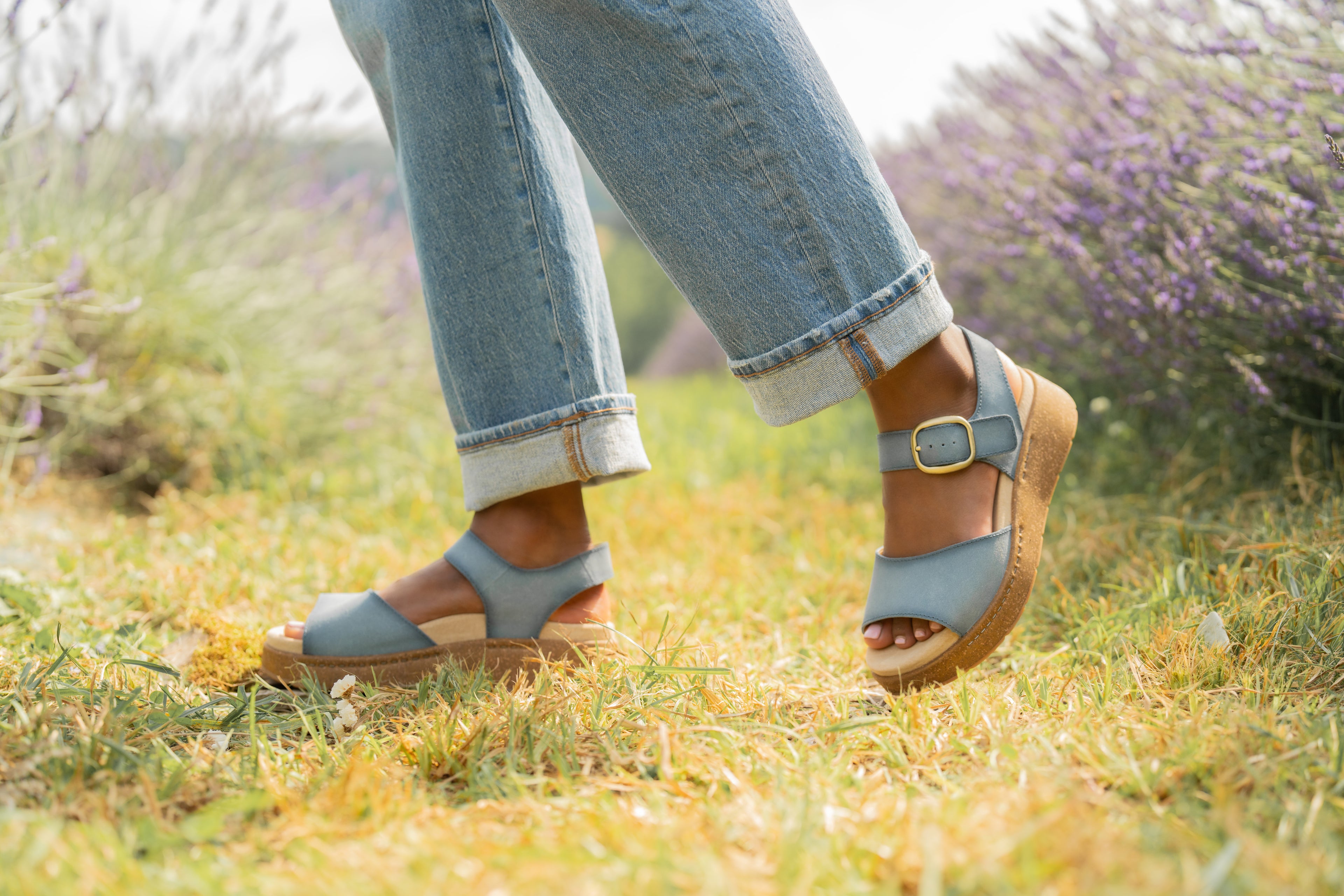 A woman walking through a grassy field with lavender bushes in the background. She is wearing Bonita Bianca Blue Dansko shoes, a sandal with dusty blue straps, gold hardware, and a thick sole.