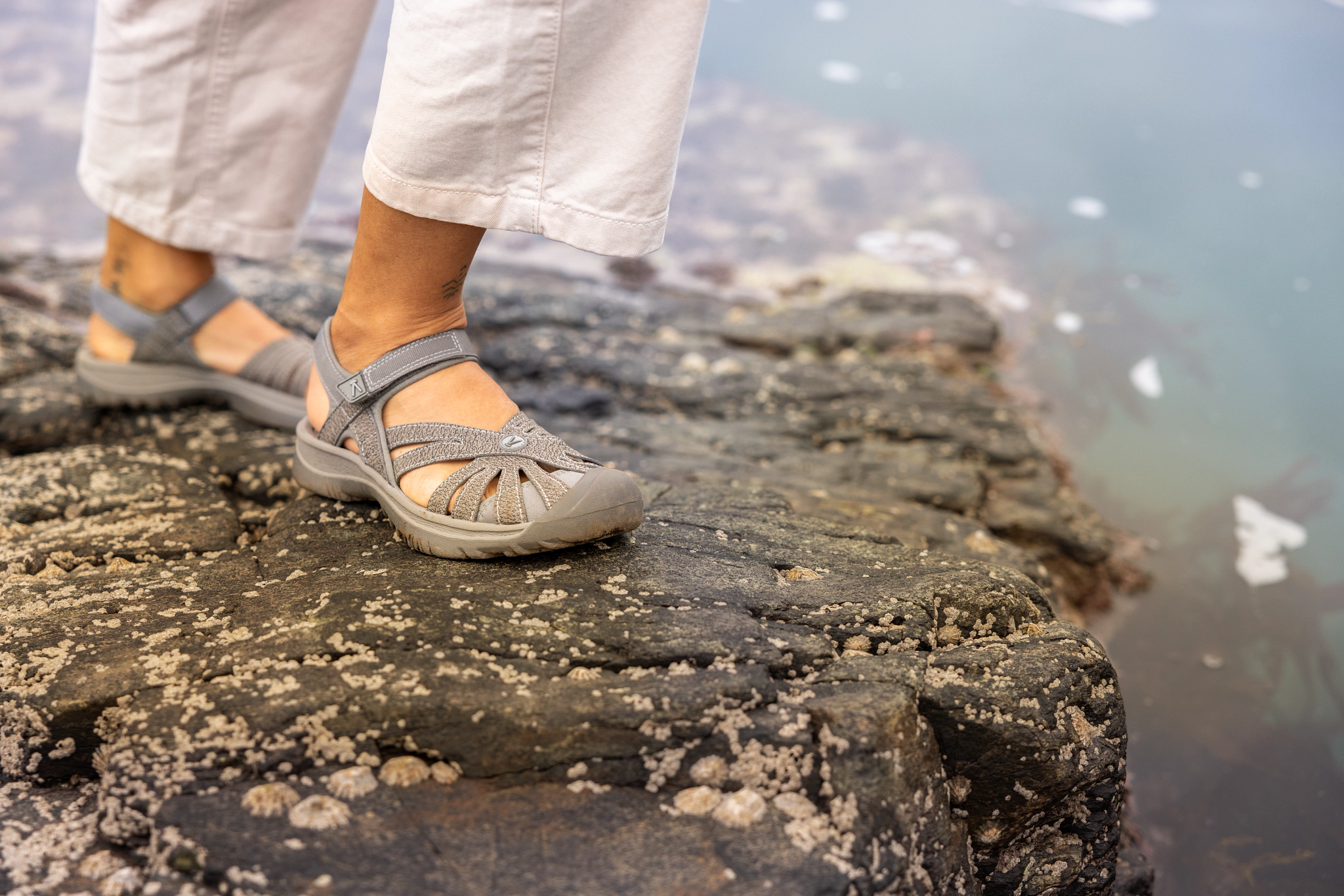 A person standing on a rocky beach shoreline wearing walking sandals.